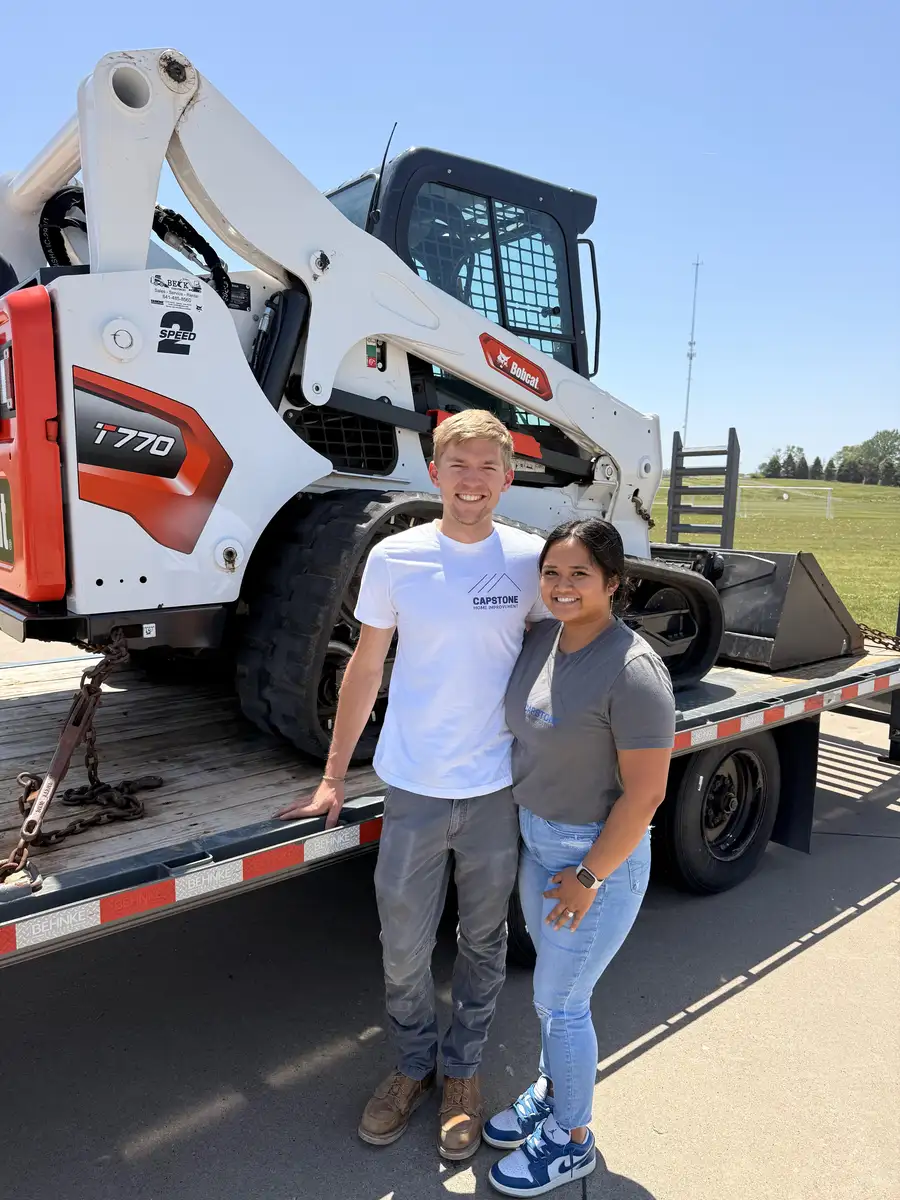 Two people standing on a trailer in front of the Capstone Bobcat T770 skid steer